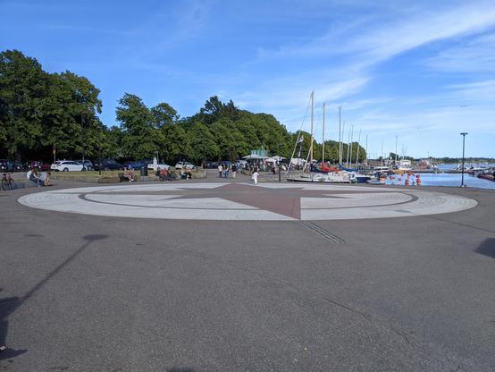 Une rose des vents au sol. Au fond à droite, on voit des bateaux amarrés. Au fond à gauche, des arbres. Le ciel est bleu, légèrement voilé.