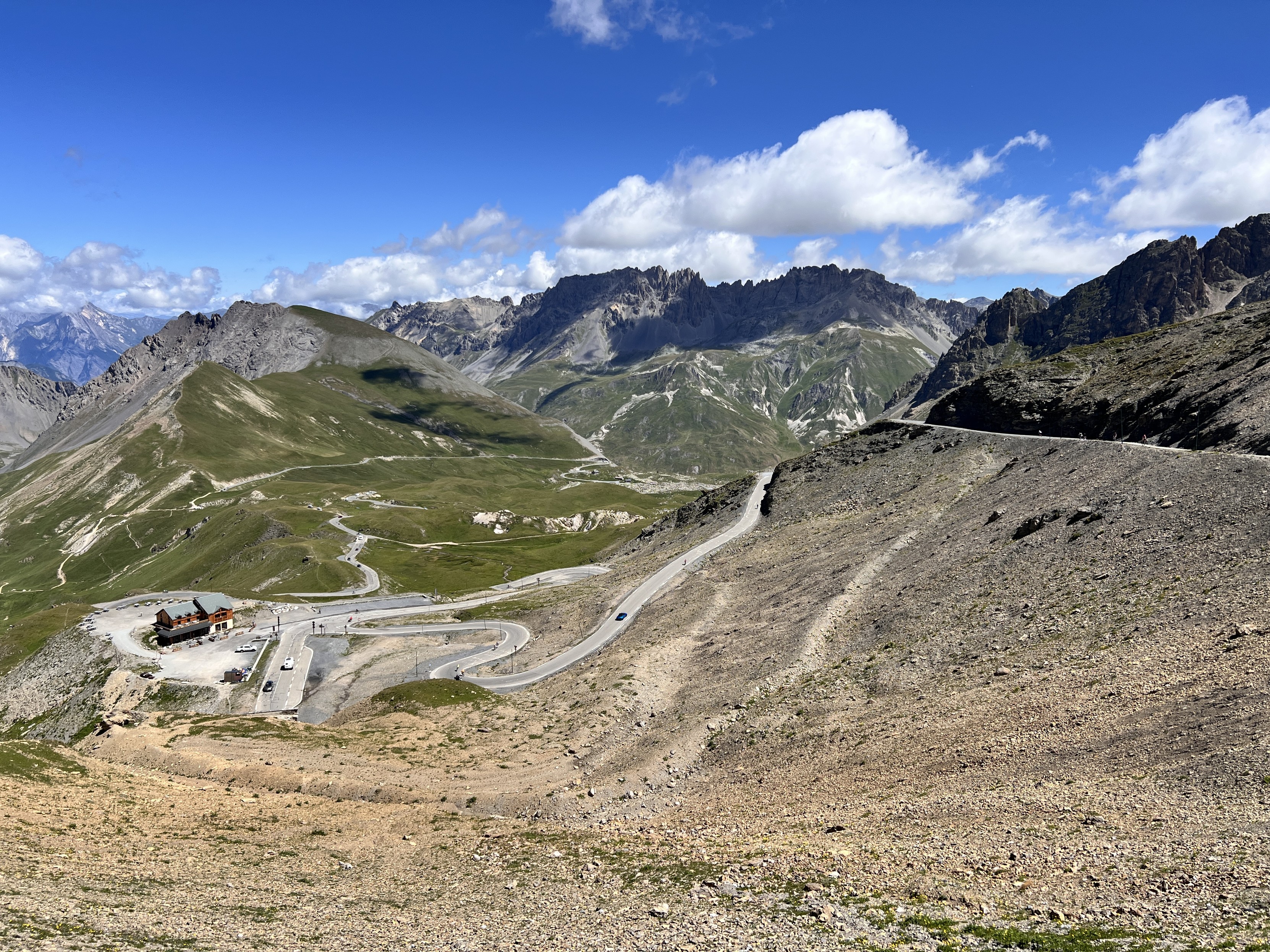 Paysage de montagne avec une route qui tournicote. Pas de végétation à cette altitude. En contrebas, un bâtiment avec un parking. Sur la route qui monte, de valeureux cyclistes.