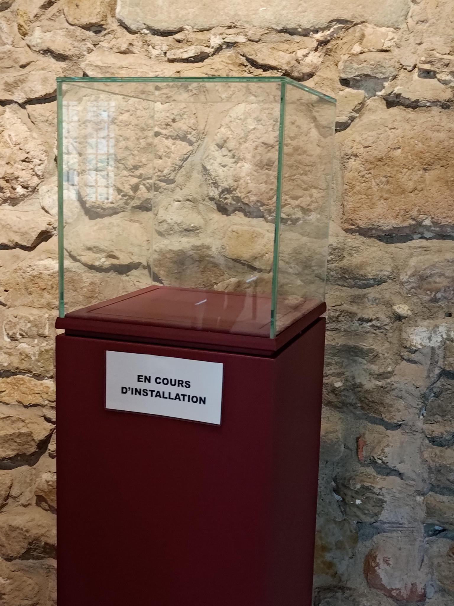 Un présentoir en bois surplombé d'une cube en verre devant un mur en pierre. Mais ce présentoir est parfaitement vide. Une écriteau sur le présentoir indique "En cours d'installation"