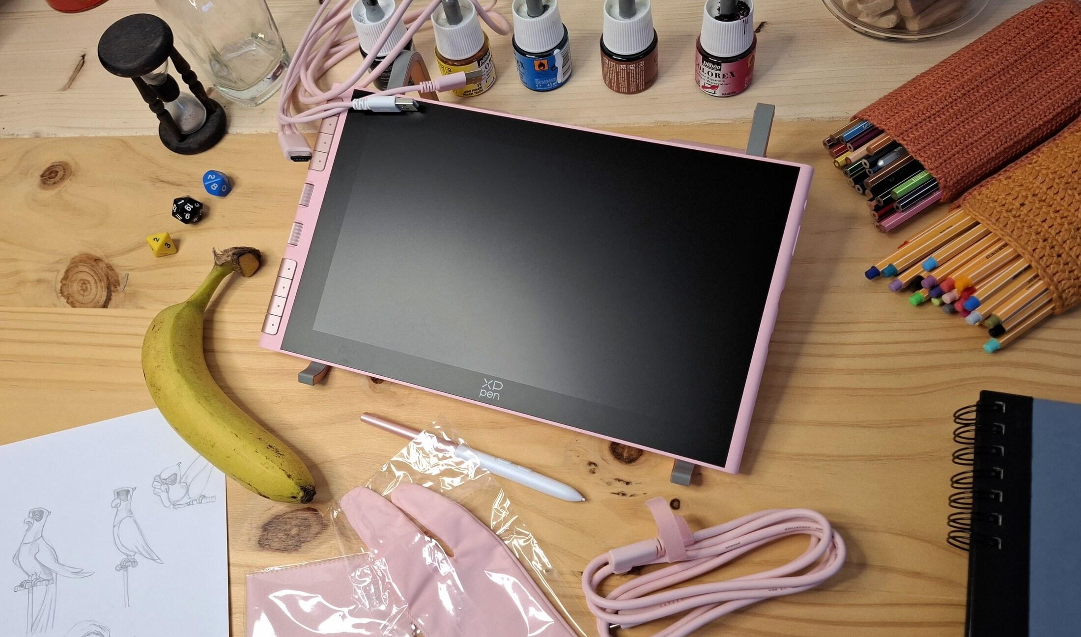 Photo of a small 12 inch drawing tablet disconnected, with all accessories around it, just after being unboxed. The tablet is pink, and so are the accessories: cables, gloves, and the stylus. On my workbench, there’s also decorative objects I use in the review: a banana, dice, colored pencils and ink set.