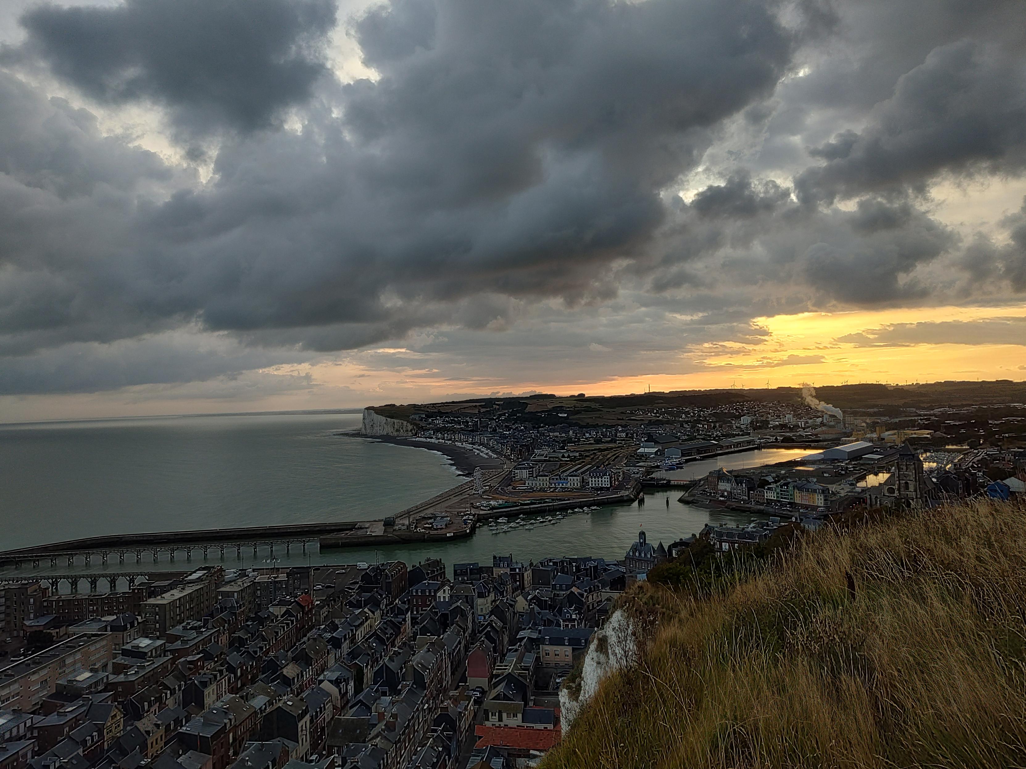 Le lever du soleil sur deux villages de bord de mer séparés par un port, au loin on voit la falaise. La photo est prise sur la falaise en face et donc en hauteur de la ville. Le ciel est nuageux mais on voit le soleil en dessous qui se lève