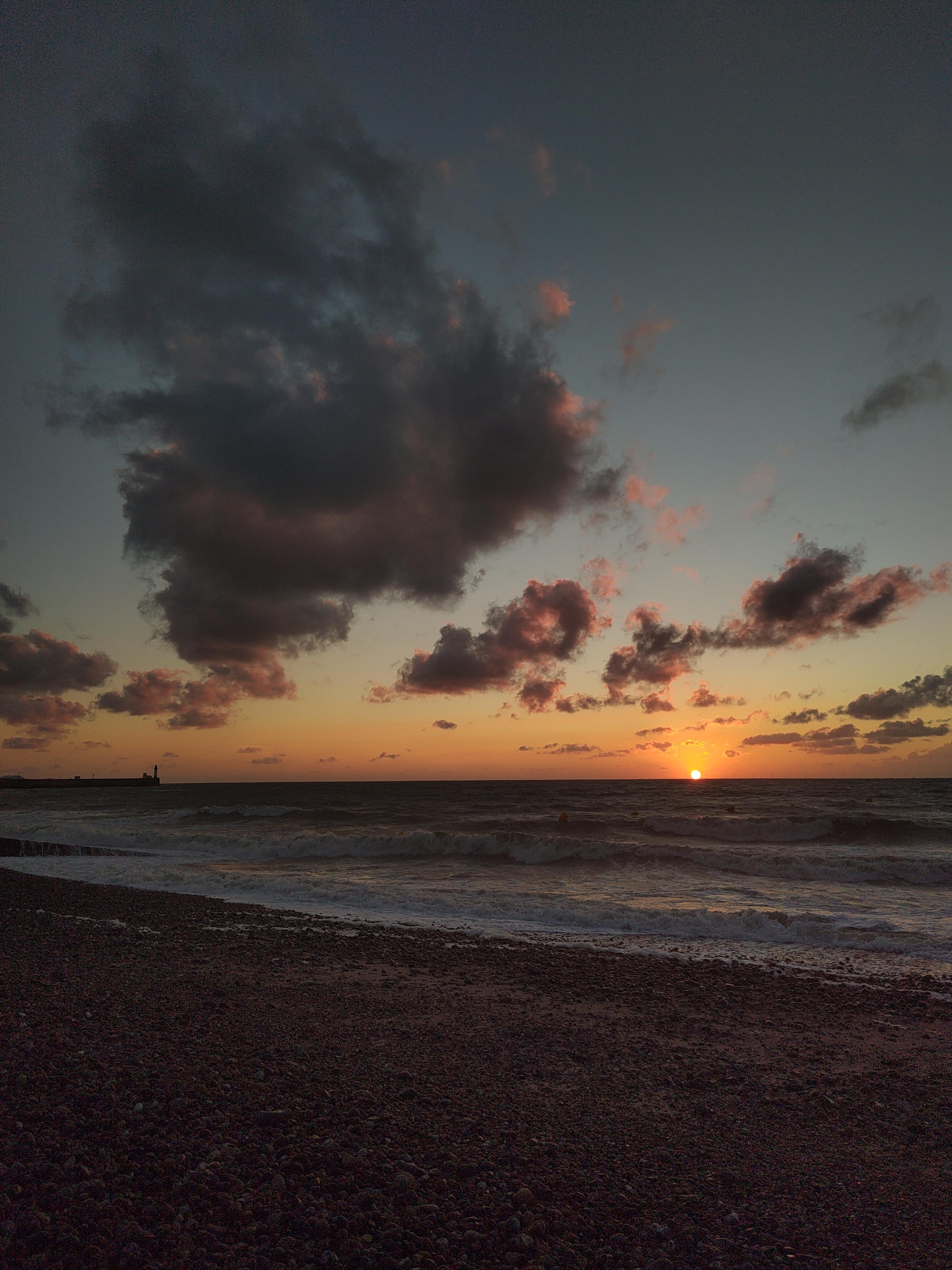 Le soleil illumine le ciel de couleur orange, alors qu'il a presque disparu derrière l'horizon, il reste quelque nuage dans le ciel. En avant plan on voit la mer et la plage de galet